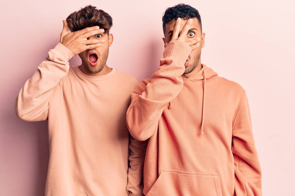 Young gay couple wearing casual clothes peeking in shock covering face and eyes with hand, looking through fingers with embarrassed expression.  - Photo, image