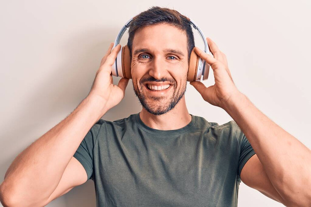 Молодий красивий чоловік слухає музику за допомогою навушників над ізольованим білим тлом, виглядає позитивно і щасливо стоячи і посміхаючись з впевненою посмішкою, показуючи зуби
 - Фото, зображення
