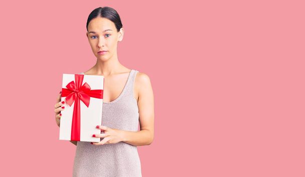 Beautiful brunette young woman holding gift thinking attitude and sober expression looking self confident  - Photo, Image