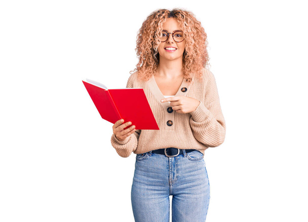 Young blonde woman with curly hair wearing glasses and reading book smiling happy pointing with hand and finger  - Zdjęcie, obraz