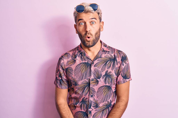 Handsome blond man on vacation wearing casual shirt and sunglasses over pink background afraid and shocked with surprise expression, fear and excited face. - Photo, Image