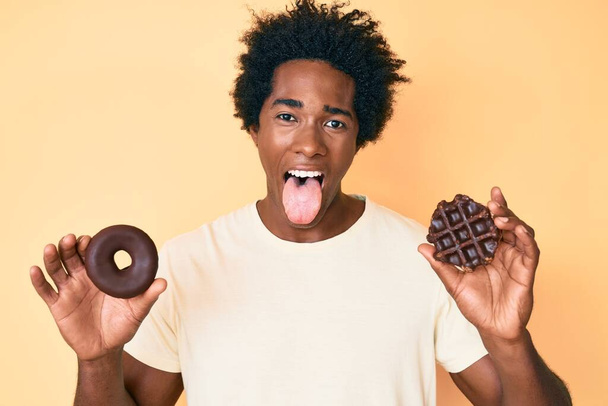 Нахабний афроамериканець з волоссям афроамериканця, який тримає шоколадний пончик і вафель, що тримає язик щасливим смішним виразом..  - Фото, зображення