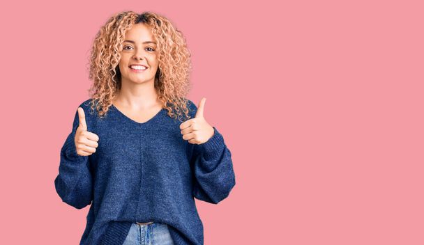 Young blonde woman with curly hair wearing casual winter sweater success sign doing positive gesture with hand, thumbs up smiling and happy. cheerful expression and winner gesture.  - Photo, Image
