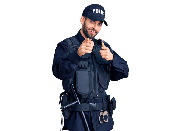 Młody, przystojny mężczyzna w brodzie, w mundurze policyjnym, wytykający palcami kamerę ze szczęśliwą i zabawną twarzą. dobra energia i wibracje.  - Zdjęcie, obraz
