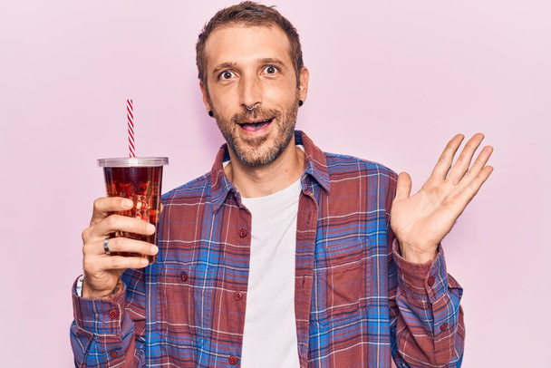 Young handsome man drinking glass of cola beverage celebrating achievement with happy smile and winner expression with raised hand  - Foto, Bild
