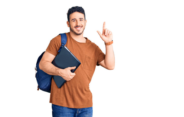 Joven hombre hispano con mochila estudiantil sosteniendo aglutinante sorprendido con una idea o pregunta apuntando con el dedo con cara feliz, número uno  - Foto, imagen
