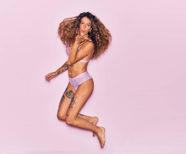 Young beautiful curly woman with tattoo wearing lingerie smiling happy. Jumping with smile on face throwing kiss with hands over isolated pink background - Foto, imagen