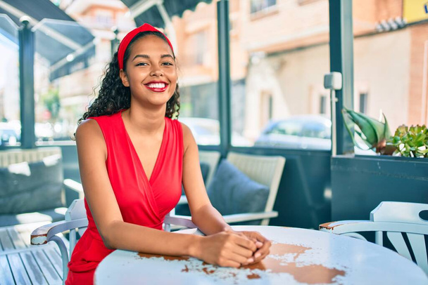 Jovem afro-americana sorrindo feliz sentando-se na mesa no terraço da loja de café. - Foto, Imagem