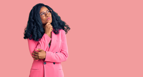 Όμορφη Αφροαμερικανή γυναίκα που φοράει επαγγελματικό σακάκι και γυαλιά σοβαρό πρόσωπο σκέφτεται την ερώτηση με το χέρι στο πηγούνι, στοχαστική για σύγχυση ιδέα  - Φωτογραφία, εικόνα