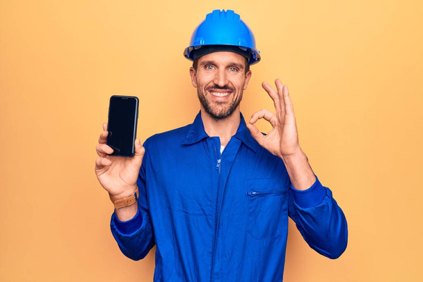 Mladý pohledný dělník v uniformě a klobouku drží smartphone ukazující obrazovku dělá v pořádku znamení s prsty, usmívá přátelské gesto vynikající symbol - Fotografie, Obrázek