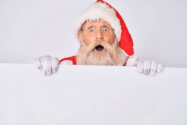 Vanha vanhempi mies harmaat hiukset ja pitkä parta yllään joulupukki puku tilalla banneri peloissaan ja järkyttynyt yllätys ja hämmästynyt ilme, pelko ja innoissaan kasvot.  - Valokuva, kuva