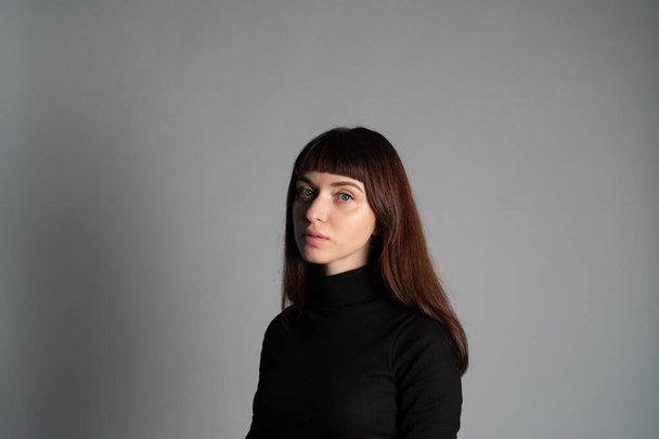Студийный портрет красивой брюнетки в сложенном черном свитере с поло-шеей, смотрящей в камеру, на сером фоне - Фото, изображение