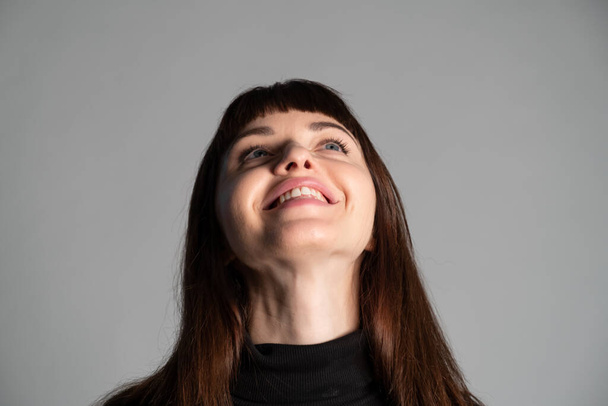Großaufnahme des Studioporträts einer hübschen brünetten Frau, die einen gefalteten schwarzen Polohals-Pullover trägt, lacht und zur Seite schaut, vor einem schlichten grauen Hintergrund - Foto, Bild