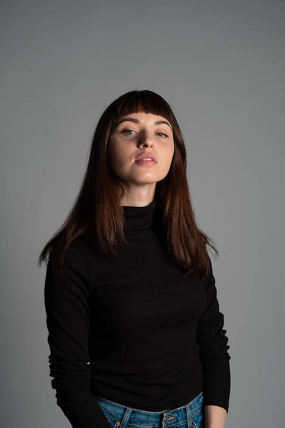 Studio portrait of a pretty brunette woman, wearing folded black polo-neck sweater, looking at camera, against a plain grey background - Foto, afbeelding