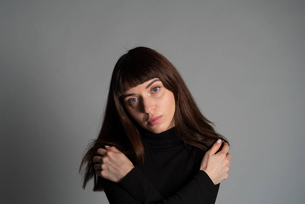 Nahaufnahme des Studioporträts einer hübschen brünetten Frau, die einen gefalteten schwarzen Polohals-Pullover trägt, die Hände auf den Schultern, in die Kamera blickt, vor einem schlichten grauen Hintergrund - Foto, Bild