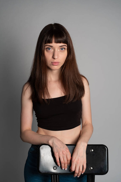 Studio portret ładnej brunetki w czarnej kamizelce, stojącej za krzesłem, poważnie patrzącej w kamerę, na szarym tle - Zdjęcie, obraz