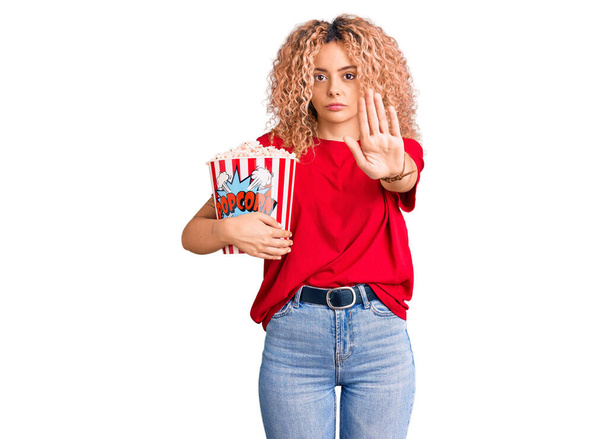 Mujer rubia joven con pelo rizado comiendo palomitas de maíz con la mano abierta haciendo stop sign con expresión seria y segura, gesto de defensa  - Foto, Imagen