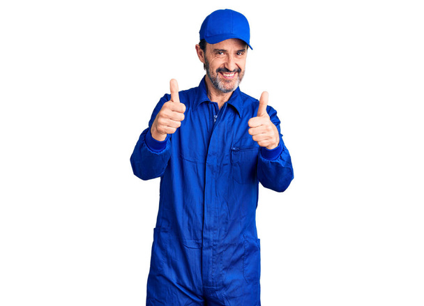 Gut aussehender Mann mittleren Alters in Mechaniker-Uniform mit positiver Geste mit der Hand, Daumen hoch lächelnd und glücklich. Fröhlicher Ausdruck und Siegergeste.  - Foto, Bild