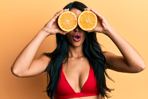 Mulher hispânica bonita usando biquíni segurando laranja nos olhos com medo e chocado com a expressão surpresa e surpreendente, medo e rosto animado.  - Foto, Imagem