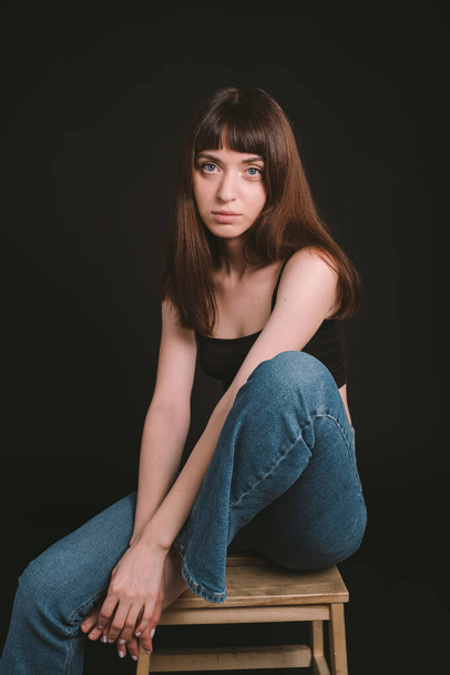 Studio portrait of a pretty barefoot brunette woman in black spaghetti strap top and jeans, looking at the camera, sitting against a plain black background - Photo, Image