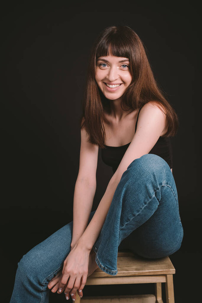 Studio portrait of a pretty barefoot brunette woman in black spaghetti strap top and jeans, looking at the camera, sitting against a plain black background - 写真・画像