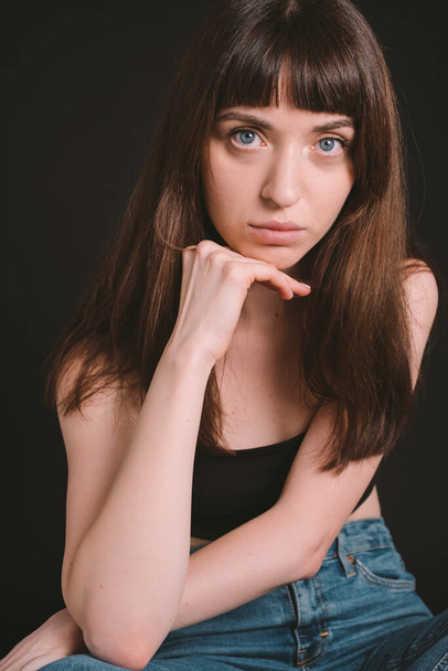 Studio portrait of a pretty brunette woman in black spaghetti strap top and jeans, looking at the camera, sitting against a plain black background - 写真・画像