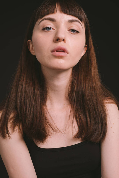 Close up studio portrait of a pretty brunette woman in a black spaghetti strap top, looking at the camera, against a plain black background - Zdjęcie, obraz