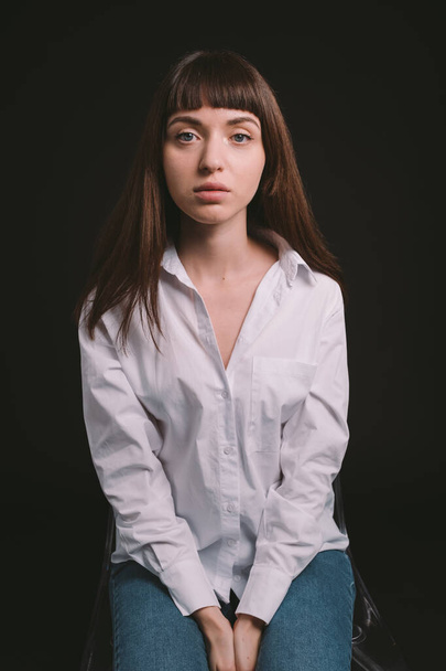 Studio portrait of a pretty brunette woman in a white shirt, sitting on a chair, against a plain black background, seriously looking at the camera - Fotografie, Obrázek