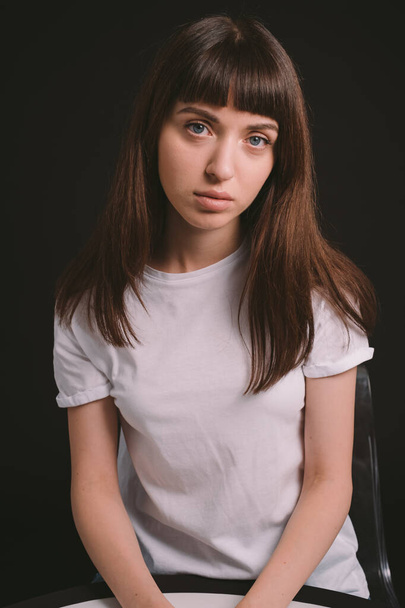 Studio portrait of a pretty brunette woman in a white blank t-shirt, sitting against a plain black background, looking at the camera - Photo, Image