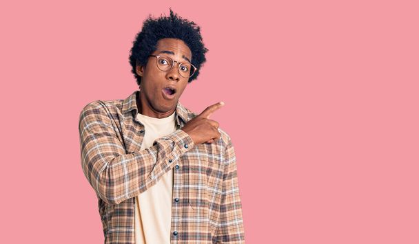 Handsome african american man with afro hair wearing casual clothes and glasses surprised pointing with finger to the side, open mouth amazed expression.  - Photo, image