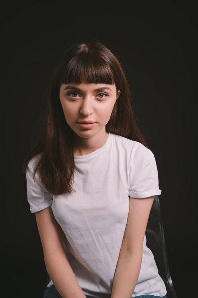 Studio portrait of a pretty brunette woman in a white blank t-shirt, against a plain black background, looking at the camera - Foto, immagini