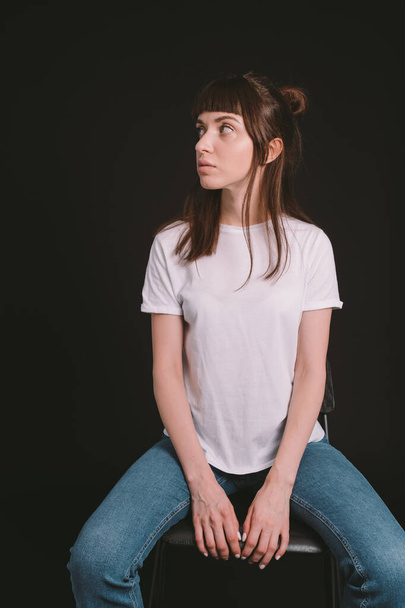 Studio portrait of a pretty brunette woman in a white blank t-shirt, sitting on a chair, against a plain black background, looking to the side - Photo, Image