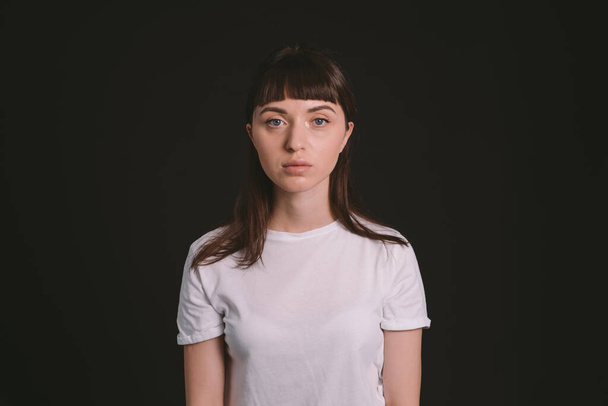Studio portrait of a pretty brunette woman in a white blank t-shirt, against a plain black background, looking at camera - Fotografie, Obrázek