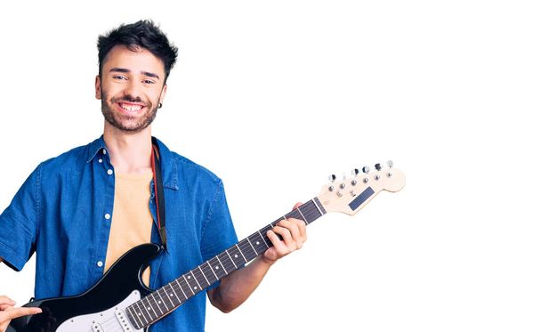 Joven hispano tocando la guitarra eléctrica sonriendo feliz señalando con la mano y el dedo  - Foto, imagen