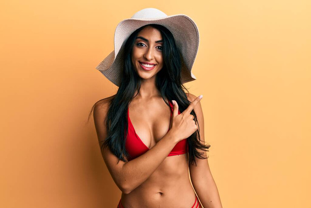 Mulher hispânica bonita usando biquíni e chapéu de verão sorrindo alegre apontando com a mão e o dedo para o lado  - Foto, Imagem