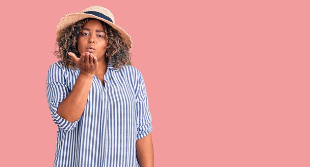 Young african american plus size woman wearing summer hat looking at the camera blowing a kiss with hand on air being lovely and sexy. love expression.  - Zdjęcie, obraz
