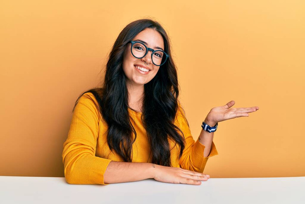 Beautiful brunette young woman wearing glasses and casual clothes sitting on the table smiling cheerful presenting and pointing with palm of hand looking at the camera.  - Foto, Imagem