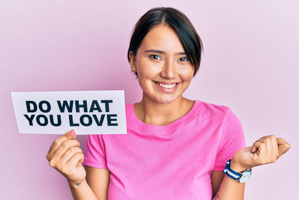 Beautiful young woman with short hair holding paper with do what you love text screaming proud, celebrating victory and success very excited with raised arm  - Photo, Image
