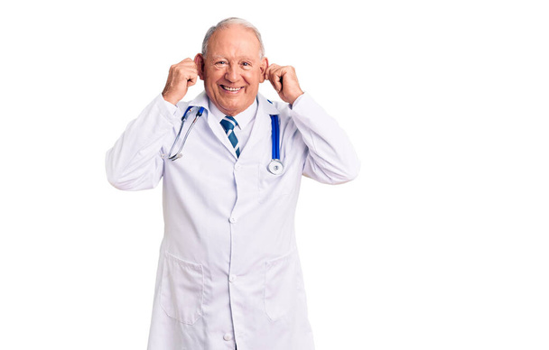 Uomo anziano con i capelli grigi che indossa cappotto medico e stetoscopio sorridente tirando le orecchie con le dita, gesto divertente. problema di audizione  - Foto, immagini
