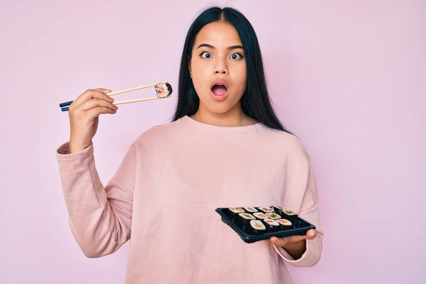 若い美しいアジアの女の子は箸を使用して寿司を食べて恐れて、驚きと驚きの表情でショックを受けました,恐怖と興奮した顔.  - 写真・画像