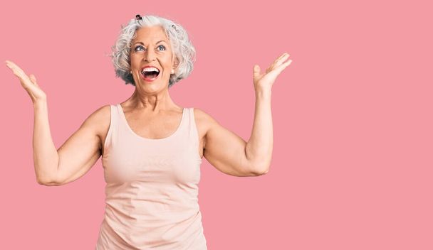 Старшая седовласая женщина в повседневной одежде празднует победу со счастливой улыбкой и выражением лица победителя с поднятыми руками  - Фото, изображение