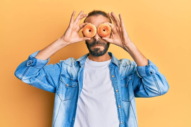 Attractive man with long hair and beard holding tasty colorful doughnuts on eyes relaxed with serious expression on face. simple and natural looking at the camera.  - Foto, imagen