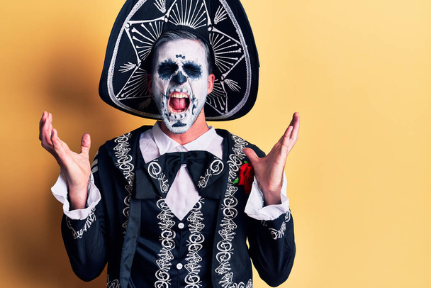 Jeune homme portant le jour mexicain du costume mort sur jaune célébrant fou et fou de succès avec les bras levés et les yeux fermés hurlant excité. concept gagnant  - Photo, image