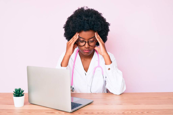 Jonge Afro-Amerikaanse vrouw draagt dokter stethoscoop werken met behulp van computer laptop die lijdt aan hoofdpijn wanhopig en gestrest omdat pijn en migraine. handen op het hoofd.  - Foto, afbeelding