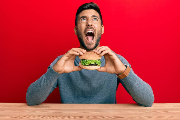 Przystojny Latynos jedzący pysznego hamburgera wściekłego i szalonego krzyczącego sfrustrowanego i wściekłego, krzyczącego ze złości patrzącego w górę.  - Zdjęcie, obraz