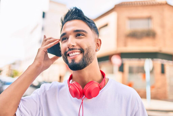 Hombre árabe joven sonriendo feliz escuchando mensaje de audio utilizando el teléfono inteligente en la ciudad. - Foto, Imagen