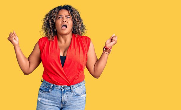 若いアフリカ系アメリカ人のプラスサイズの女性が袖のないシャツをクレイジーで狂った叫びと積極的な表現と腕を上げて叫んでカジュアルなスタイルを身に着けています。フラストレーションコンセプト.  - 写真・画像