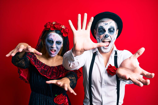 Beautiful couple wearing day of the dead costume gesturing as a zombie over isolated red background. - Photo, Image