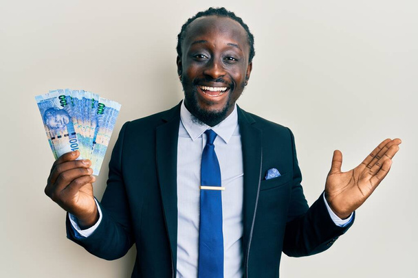 幸せな笑顔で達成を祝う100枚の紙幣を保持するビジネススーツを身に着けているハンサムな若い黒人男性と上げ手で勝者の表現  - 写真・画像