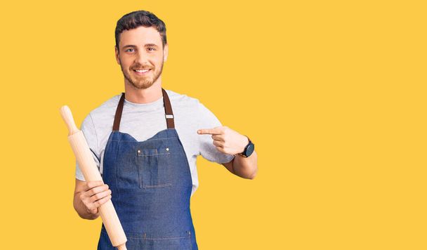 Hübscher junger Mann mit Bär in professioneller Bäckerschürze mit Knetrolle zeigt mit dem Finger auf einen selbst lächelnden glücklich und stolz  - Foto, Bild